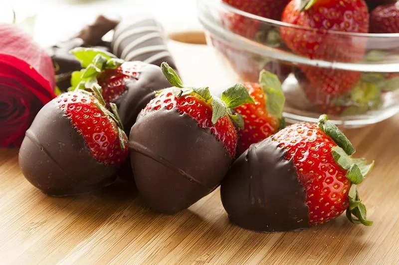 Chocolate-covered strawberries on a wooden surface with a bowl of strawberries in the background.