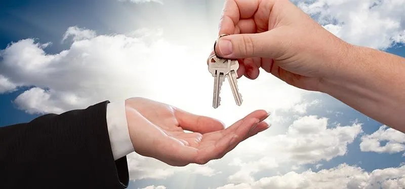 Keys being handed over between two hands with a cloudy sky background.