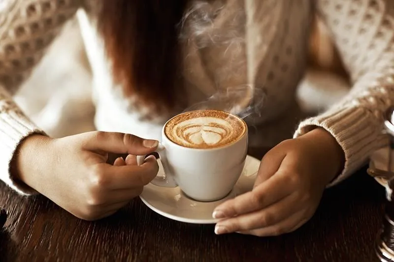 A person holding a steaming cup of coffee with latte art on a saucer.