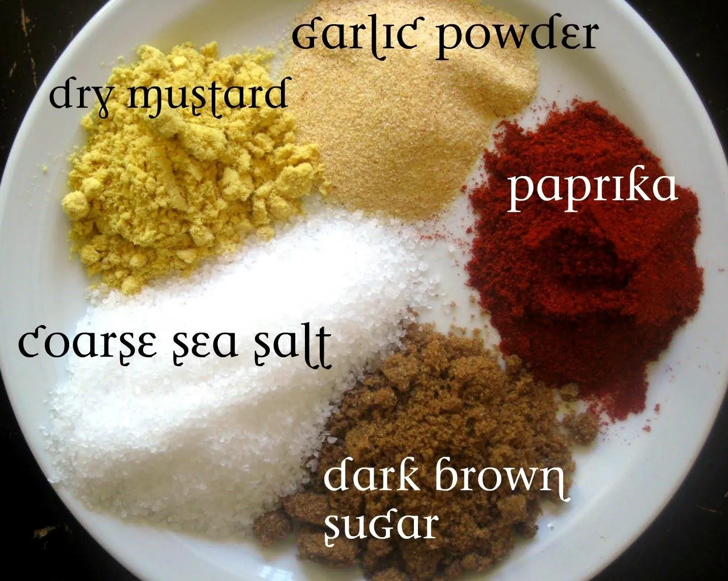A plate of various spices, including garlic powder, dry mustard, paprika, coarse sea salt, and dark brown sugar.