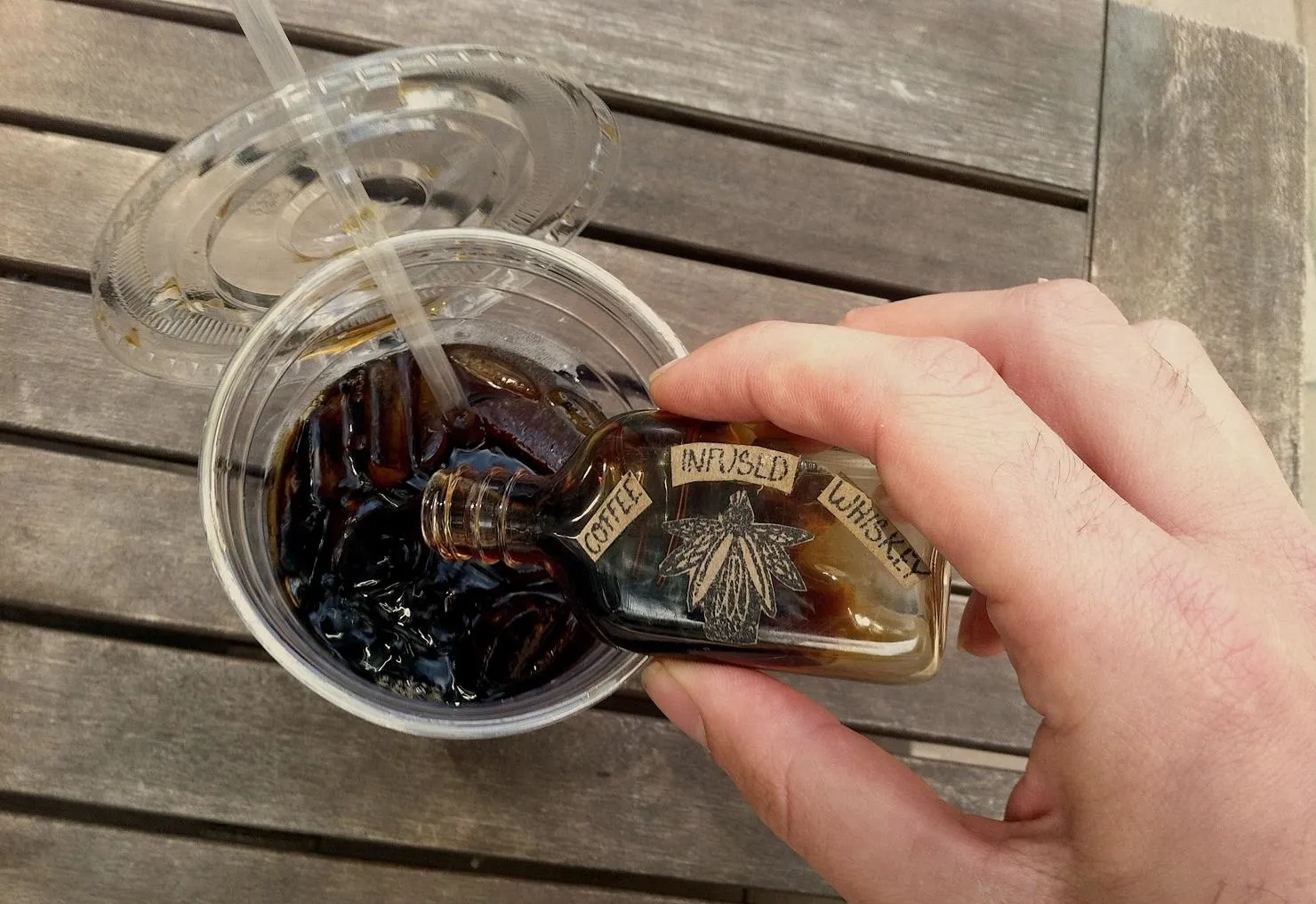A hand pouring syrup into a cup of iced coffee on a wooden table.