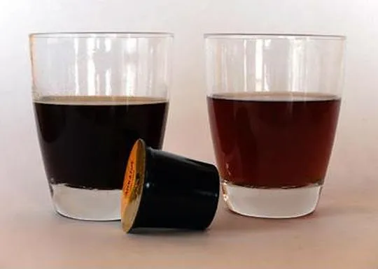Two glasses of dark and light beverages with a coffee capsule in front.
