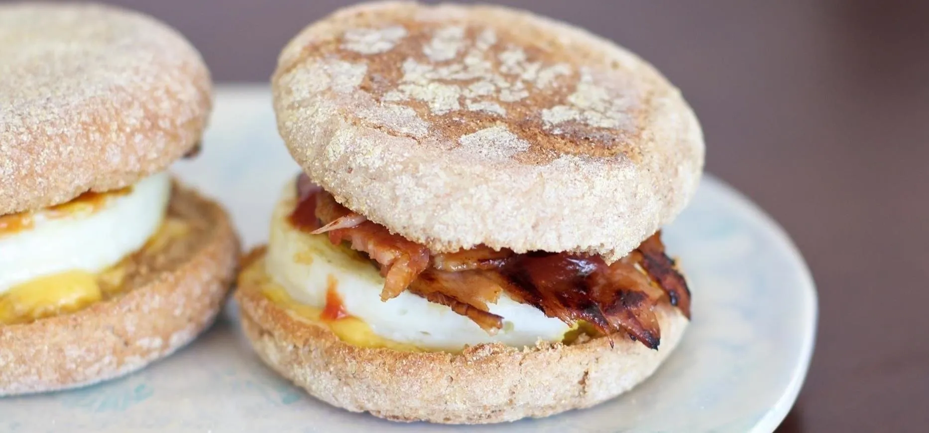 Two breakfast sandwiches on a blue-white marbled white plate, both with egg, bacon, and cheese on toasted English muffins.