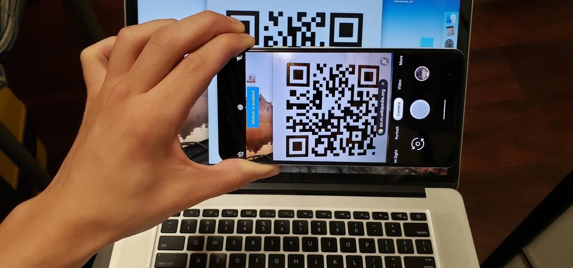 A person scanning a QR code on a smartphone while it is positioned over a laptop screen.