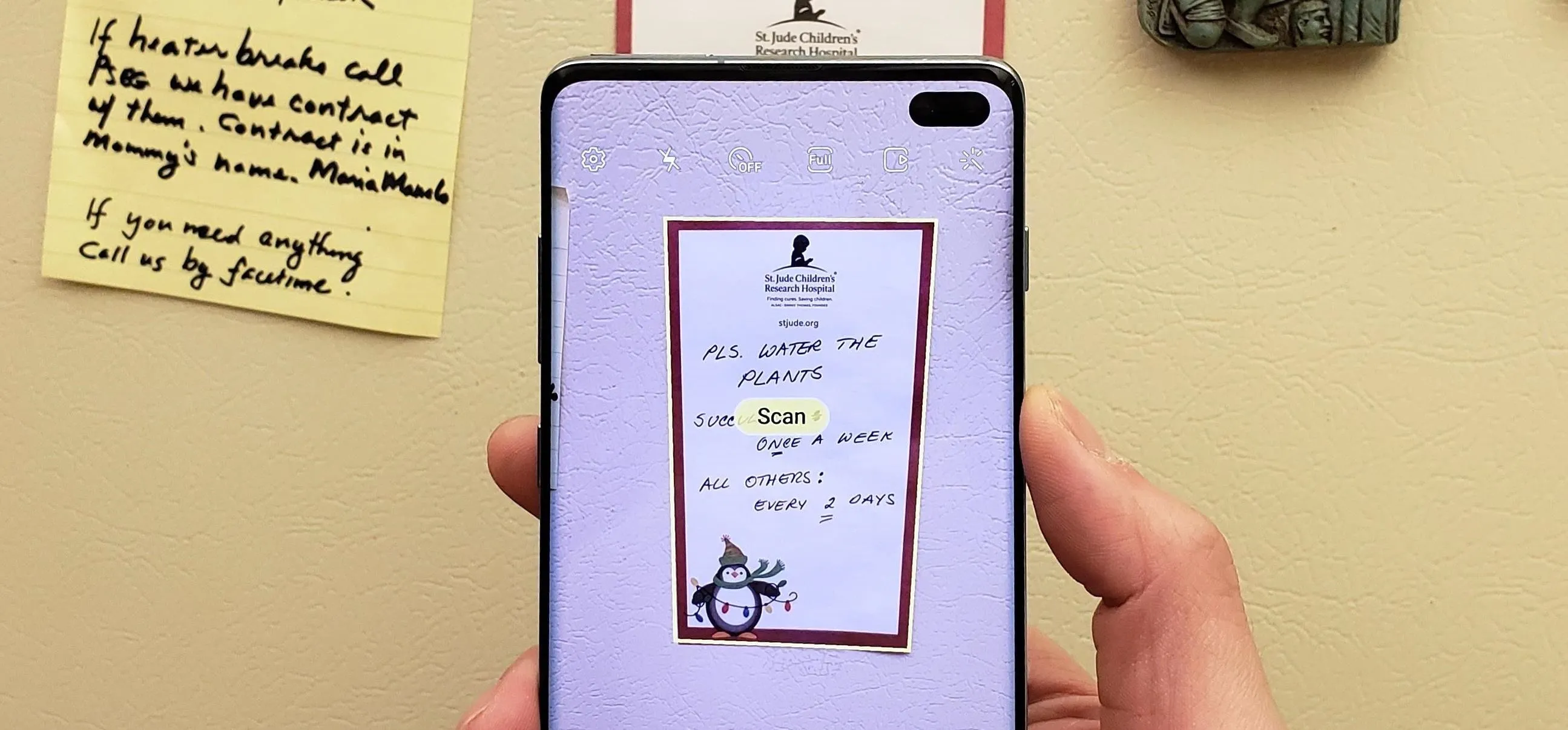 A hand holding a smartphone displaying a note on a wall with a cartoon figure at the bottom.